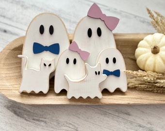 May include: A family of five white wooden ghosts with painted black eyes and blue and pink bows. The ghosts are arranged in a wooden bowl with a white cat and a white dog.