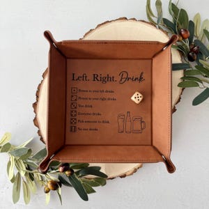 Może przedstawiać: Brązowa skórzana taca z napisem "Left. Right. Drink" i instrukcjami gry. Zawiera kostkę do gry i ilustracje szklanek do piwa. Taca jest kwadratowa z zaokrąglonymi rogami i jest umieszczona na drewnianym plastrze.