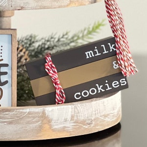 Puede incluir: Un letrero de madera marrón con el texto "milk & cookies" en letras blancas. El letrero está atado con un cordel rojo y blanco y está sentado en una bandeja de madera de varios niveles.