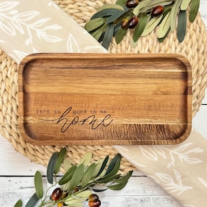 May include: A rectangular wooden serving tray with rounded edges. The tray is made of light brown wood and has the text "It's so good to be home" engraved on the surface.