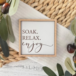May include: A wooden sign with the text "SOAK. RELAX. enjoy" in brown lettering on a white background. The sign is framed with a light brown wood frame.