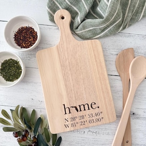 May include: A light-coloured wooden chopping board with a handle, featuring the word "home" and coordinates. Two small white bowls with spices, wooden spoons, and a green striped tea towel are also visible.