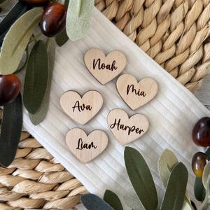 Peut inclure: Cinq marque-places en bois en forme de cœur avec des noms écrits en cursive. Les noms sont Noah, Mia, Ava, Harper et Liam.