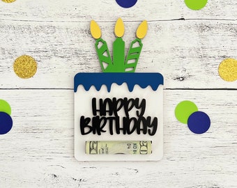 May include: A wooden birthday card shaped like a cake with green candles and blue frosting. The card has a slot for cash and says "Happy Birthday" in black letters.