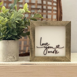 Może przedstawiać: Kwadratowy znak w drewnianej ramie z napisem "love you more" w kursywie. Znak ma białe tło i postarzaną, jasnobrązową ramę. Mała roślina doniczkowa znajduje się po lewej stronie znaku.