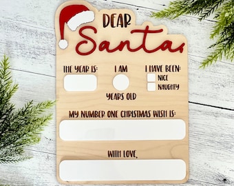 Puede incluir: Un letrero de madera con un gorro de Papá Noel en la parte superior. El letrero dice "Querido Papá Noel" y tiene espacios para que un niño escriba su edad, su deseo de Navidad y si ha sido bueno o malo.