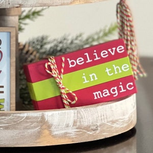 May include: A red and green wooden sign with the text "believe in the magic" tied with a red and yellow twine bow.