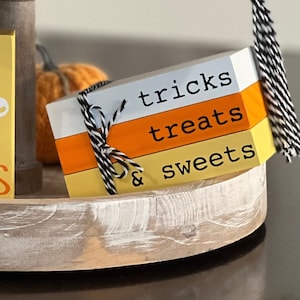 Può includere: Un cartello di legno a tre livelli con le parole "tricks treats & sweets" in lettere nere su uno sfondo bianco, arancione e giallo. Il cartello è legato con uno spago nero e bianco.