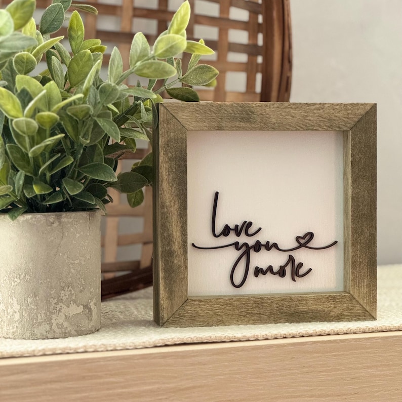 Może przedstawiać: Kwadratowy drewniany znak w ramie z napisem "Love you more" w kursywie. Znak ma białe tło i ciemnobrązową ramę. Mała roślina doniczkowa znajduje się po lewej stronie znaku.