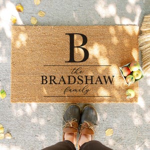 Könnte beinhalten: Rechteckige Fußmatte aus Naturkokosfaser mit dem schwarzen Buchstaben "B" über den Worten "the BRADSHAW family" in Schreibschrift. Die Matte liegt auf einer Betonoberfläche, im Vordergrund ein Paar braune und graue Schuhe. Äpfel in einem Behälter.