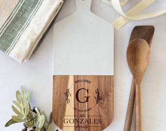 May include: A personalized cutting board with a white marble top and a wooden base. The wooden base is engraved with the words "WELCOME TO OUR HOME" and "THE GONZALES FAMILY" around a large letter "G". Two wooden spoons and a green and white striped towel are next to the board.