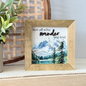 May include: Square wooden framed art print with a watercolor mountain landscape scene. The print features a lake, evergreen trees, and snow-capped mountains. The text "not all who wander are lost" is displayed on the print.