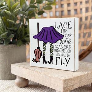 Puede incluir: Un letrero cuadrado de madera blanca con un gráfico de una falda morada, medias a rayas verdes y blancas, botas negras y una escoba marrón. El texto dice: LACE UP YOUR BOOTS GRAB YOUR BROOMSTICK IT'S TIME TO FLY en letras negras.