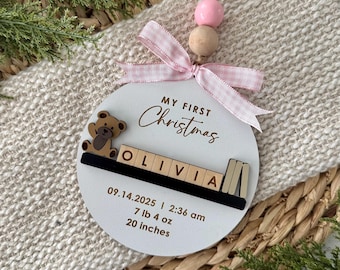 May include: A white, round Christmas ornament with the text "MY FIRST Christmas" and the name "OLIVIA". It features a teddy bear, Scrabble-style letters, and a pink ribbon. The ornament includes the date 09.14.2025, time, weight, and height (50.8 cm).