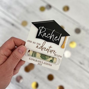 May include: A white card with a black graduation cap and the name "Rachel" written in cursive. The card says "And so the adventure begins" and "Congratulations 2023". A rolled-up dollar bill is visible in the card.