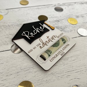 May include: A white wooden card with a black graduation cap and the name "Rachel" written in white. The card says "And so the adventure begins" and has a rolled up dollar bill inside. The card also says "Congratulations 2023".
