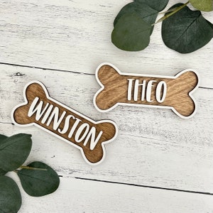 May include: Two wooden dog bone-shaped signs with white painted edges. The signs have the names "Winston" and "Theo" written in white paint.