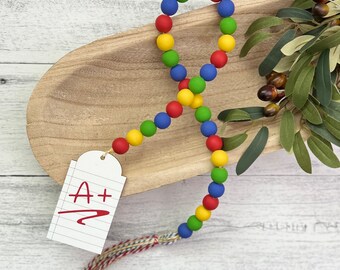 Puede incluir: Una guirnalda de cuentas de colores con cuentas rojas, amarillas, azules y verdes. La guirnalda tiene un borla en el extremo y una etiqueta blanca con el texto "A+" escrito en rojo.