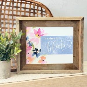 Puede incluir: Un marco de madera rústica y clara enmarca una impresión floral en acuarela en tonos rosa, azul y melocotón. La impresión presenta la frase "it's time to bloom" en escritura blanca sobre un fondo azul.