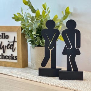 May include: Black metal restroom signs, one depicting a person and the other a person in a skirt. The figures are on rectangular bases. A framed sign with the words "hello sweet cheeks" is in the background.