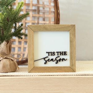 May include: A wooden framed sign with the text "'TIS THE Season" in black script. The sign has a white background and a natural wood frame. A small Christmas tree and a woven basket are in the background.