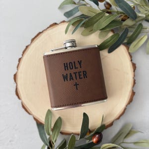 Könnte beinhalten: Eine braune Lederflasche mit den Worten "HOLY WATER" und einem Kreuz auf der Vorderseite. Die Flasche hat einen silbernen Verschluss und liegt auf einer Holzscheibe mit grünen Blättern.