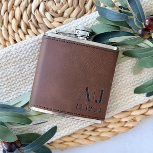 Puede incluir: Una petaca de cuero marrón con tapa y borde plateados. La petaca tiene las iniciales "AJ" y la fecha "12.12.24" grabadas. La petaca rectangular parece ser de cuero.