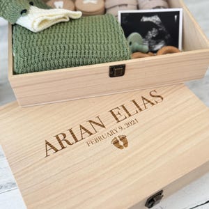 May include: A wooden keepsake box with a light wood finish. The box is engraved with the name "ARIAN ELIAS" and the date "FEBRUARY 9, 2021". The box is open and shows a green knitted blanket, a pair of white socks, and a small ultrasound image.