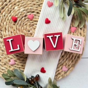 May include: Wooden blocks spelling "LOVE" in white letters and a white heart. The blocks are red and pink, with a woven background and scattered heart-shaped decorations. Green leaves and berries add a natural touch to the Valentine's Day theme.
