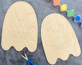May include: Two unfinished wooden ghost ornaments with the names Noah and Emily painted on them. Each ghost is holding a pumpkin. The ghosts are surrounded by paint pots and paintbrushes.