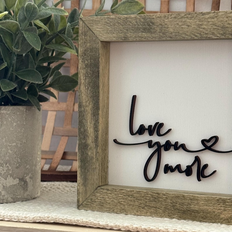 Może przedstawiać: Oprawiony drewniany znak z napisem "Love you more" w kursywie. Znak ma białe tło i rustykalną, zwietrzałą drewnianą ramę. Po lewej stronie widoczna jest mała roślina doniczkowa.