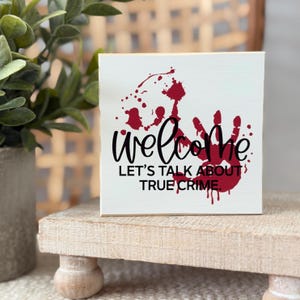 Puede incluir: Un letrero cuadrado de madera blanca con un gráfico de huella de mano roja y salpicaduras de pintura. El texto en fuente negra cursiva y sans-serif dice: welcome LET'S TALK ABOUT TRUE CRIME. El letrero tiene un acabado liso y bordes rectos.