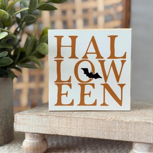 Puede incluir: Un letrero cuadrado de madera blanca con el texto naranja HALLOWEEN en una fuente serif negrita. La letra O en la palabra LOW está reemplazada por una pequeña silueta negra de un murciélago volando. El letrero tiene un acabado liso.