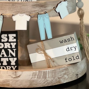 May include: A tiered wooden tray with a laundry theme. The top tier has a string of blue and white clothes cutouts, including a t-shirt and jeans. The bottom tier has a stack of three wooden blocks with the words "wash", "dry", and "fold" written on them.