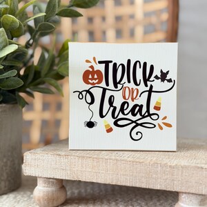 Puede incluir: Un letrero cuadrado de madera blanca con el texto negro Trick or Treat, que incluye una pequeña calabaza naranja, una araña negra, un murciélago negro y dos dulces de maíz de colores naranja, amarillo y blanco impresos en la superficie frontal.