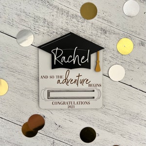 May include: White card with a black graduation cap and the name "Rachel" written in white. The card says "And so the adventure begins" and "Congratulations 2023".