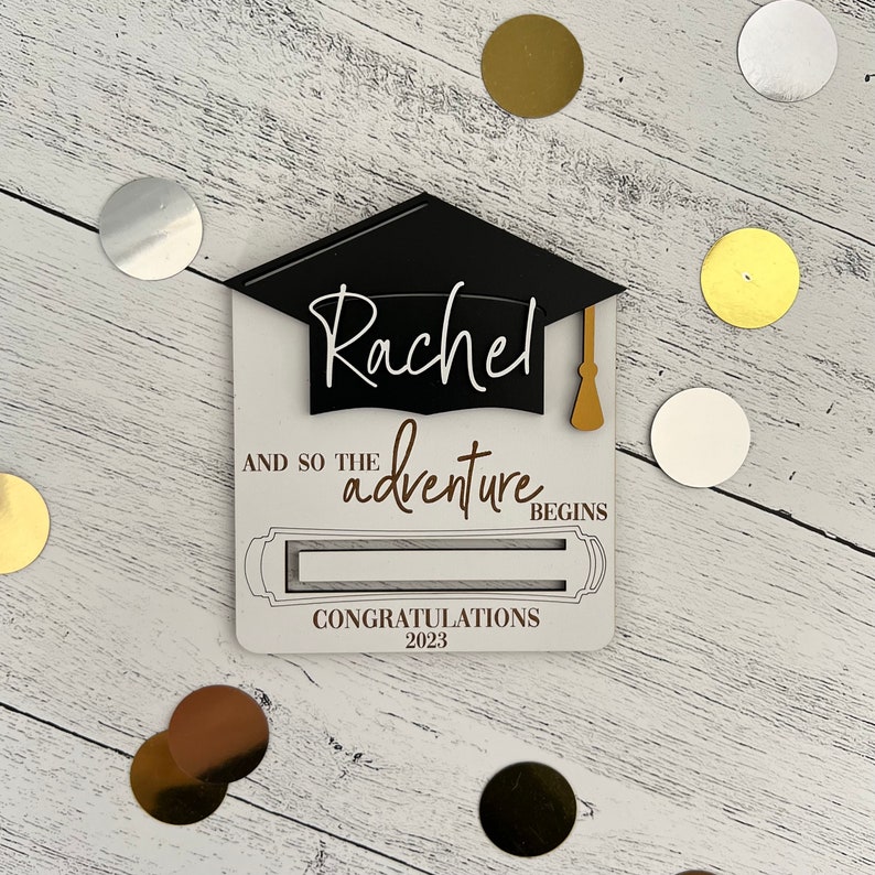 May include: White card with a black graduation cap and the name "Rachel" written in white. The card says "And so the adventure begins" and "Congratulations 2023".