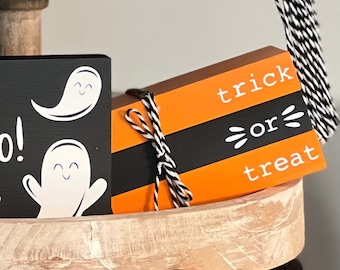 May include: A tiered wooden tray with a black and orange sign that reads "trick or treat". The sign is tied with a black and white twine. The tray is decorated with two white ghost figures on a black background.