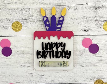 May include: A white, square birthday cake-shaped gift card holder with the words "HAPPY BIRTHDAY" in black. The cake has a pink top with three purple candles topped with gold flames. A twenty-dollar bill is visible in the slot.