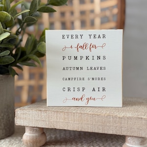 Puede incluir: Un letrero cuadrado de madera blanca con texto en negro y color óxido que dice: EVERY YEAR I fall for PUMPKINS AUTUMN LEAVES CAMPFIRE S'MORES CRISP AIR and you. El letrero tiene un acabado liso y se exhibe sobre una pequeña base de madera.