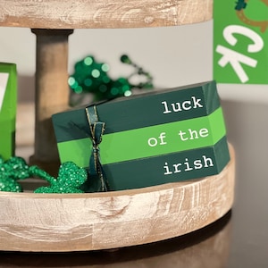 May include: A green wooden St. Patrick's Day decoration with the text "luck of the irish" in white. The blocks are tied with a green and gold ribbon. It sits on a tiered wooden tray with green shamrock accents.