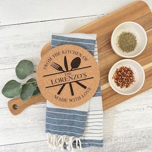 Puede incluir: Posavasos redondo de corcho con el texto "From the kitchen of The Lorenzo's Made with love" y un gráfico de un tenedor, un cuchillo y una cuchara. El posavasos está sobre una toalla de rayas azules y blancas.