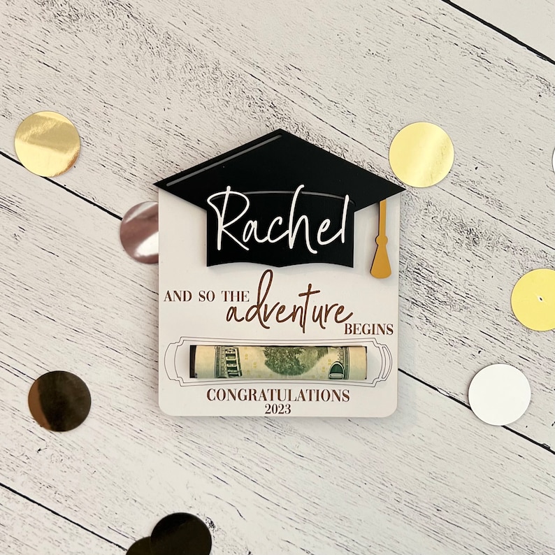 May include: A white card with a black graduation cap and the name "Rachel" written in cursive. The card says "And so the adventure begins" and "Congratulations 2023". A rolled-up dollar bill is visible in the card.