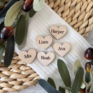 Könnte beinhalten: Fünf herzförmige Platzkarte aus Holz mit den Namen Harper, Liam, Ava, Mia und Noah. Die Platzkarte liegen auf einer weißen Oberfläche mit einer geflochtenen Strohmatte und grünen Blättern.