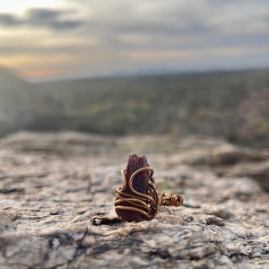 May include: A handmade ring featuring a raw, dark red gemstone wrapped in gold-colored wire. The ring is set against a natural, textured stone surface with a blurred background of a landscape and cloudy sky.