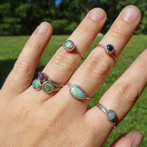 May include: A hand wearing five silver rings with different colored gemstones. The rings feature turquoise, purple, black, and white stones.