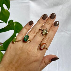 May include: A hand displays five gemstone rings. Each ring features a different colored stone, including green, orange, and clear, set in a gold-colored wire frame. The rings are displayed against a white background with a green plant.