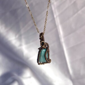 K&ouml;nnte beinhalten: Eine handgefertigte Halskette mit einem hellblauen Edelsteinanh&auml;nger, der in Kupferdraht gewickelt ist. Der Anh&auml;nger h&auml;ngt an einer zarten goldfarbenen Kette. Die Drahtwicklung bildet ein kompliziertes k&auml;figartiges Design um den Stein und zeigt die nat&uuml;rliche Sch&ouml;nheit des Edelsteins.