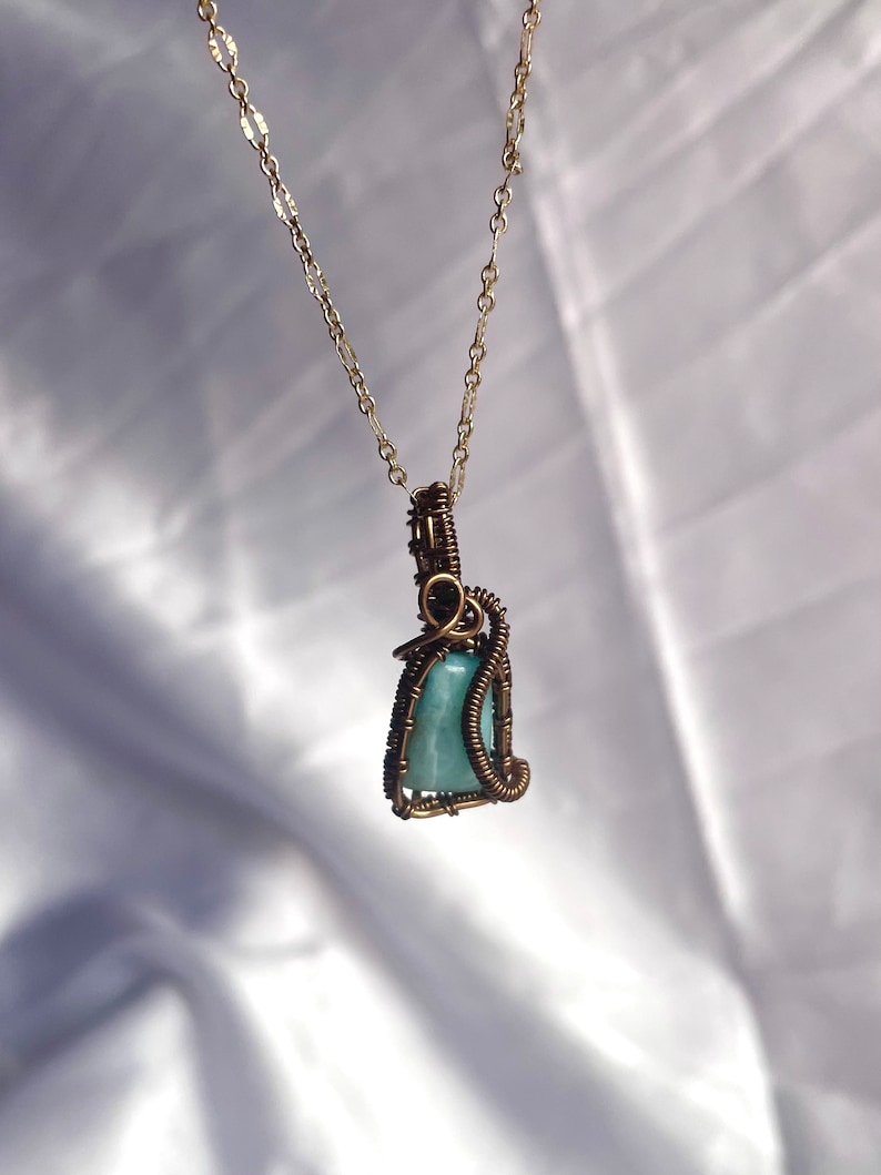 K&ouml;nnte beinhalten: Eine handgefertigte Halskette mit einem hellblauen Edelsteinanh&auml;nger, der in Kupferdraht gewickelt ist. Der Anh&auml;nger h&auml;ngt an einer zarten goldfarbenen Kette. Die Drahtwicklung bildet ein kompliziertes k&auml;figartiges Design um den Stein und zeigt die nat&uuml;rliche Sch&ouml;nheit des Edelsteins.
