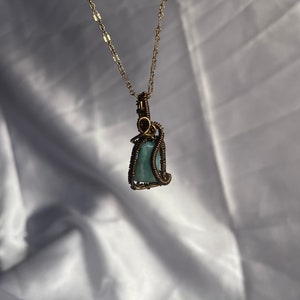 K&ouml;nnte beinhalten: Eine handgefertigte Anh&auml;ngerhalskette mit einem hellblauen Edelstein, der in bronzefarbenen Draht gewickelt ist. Der Anh&auml;nger h&auml;ngt an einer zarten goldfarbenen Kette. Die Halskette wird vor einem wei&szlig;en, strukturierten Hintergrund pr&auml;sentiert.